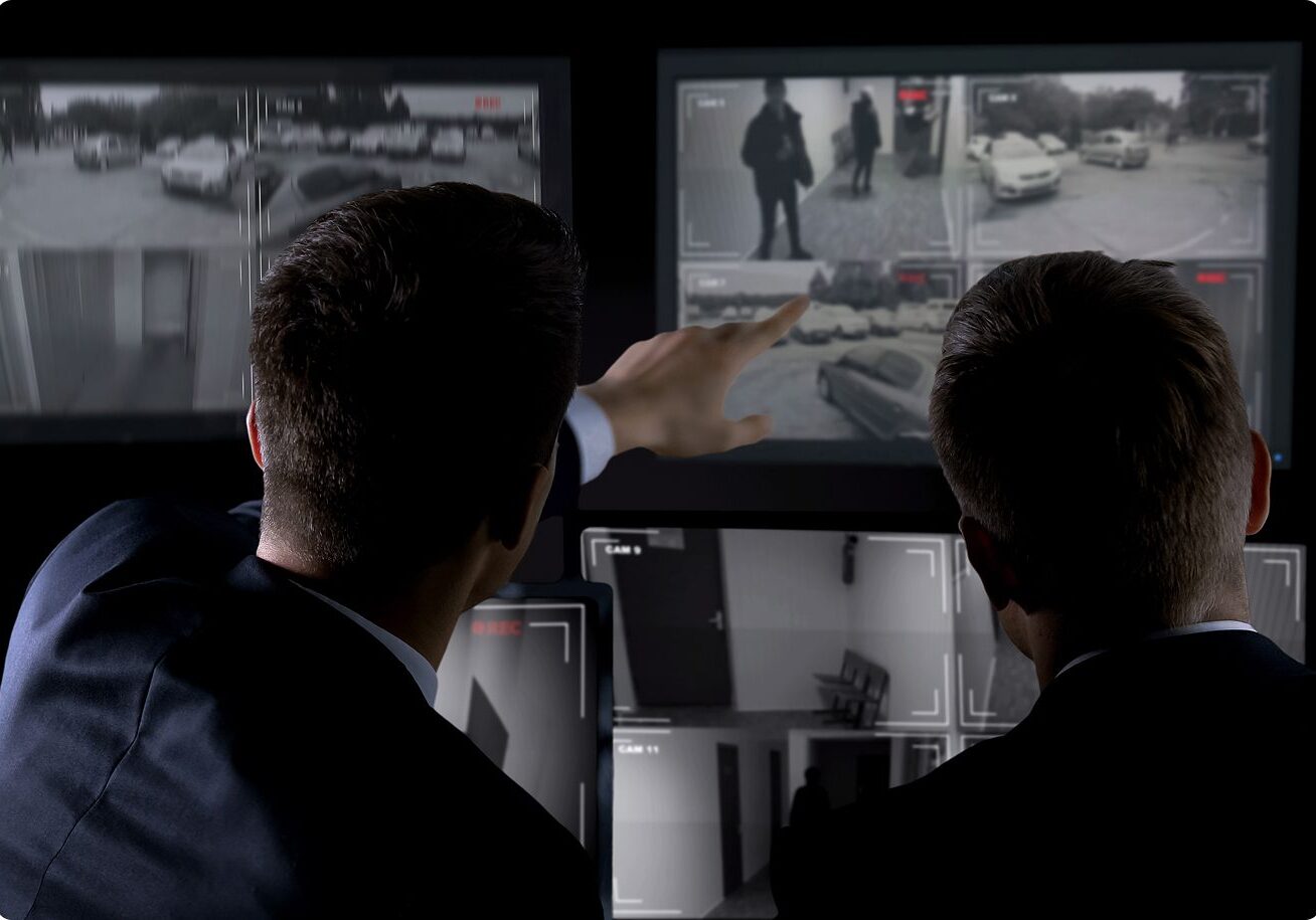 Two people in suits analyze multiple surveillance feeds on large screens, with one person pointing at one of the monitors.